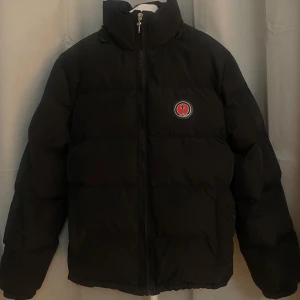 Svart pufferjacka från Trapstar - Snygg svart pufferjacka från Trapstar med dragkedja och broderad logga på bröstet. Jackan har en hög krage och texten 'Trapstar' i rött på ryggen. Perfekt för kyliga dagar.