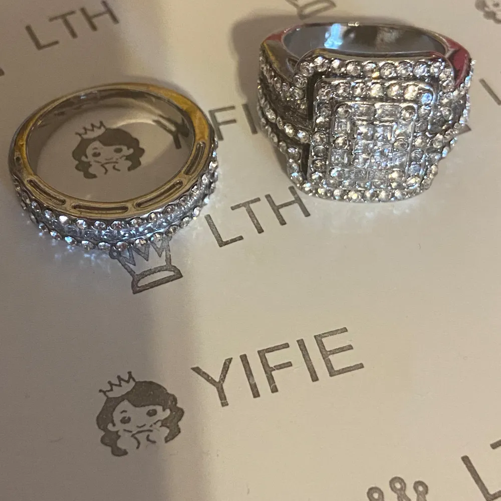 Två eleganta silverringar båda täckta med gnistrande stenar. Den ena ringen har en bredare design med en fyrkantig detalj framtill, medan den andra är smalare med en jämn rad av stenar runtom. Perfekta för att ge en lyxig touch till din stil.. Asusteet.