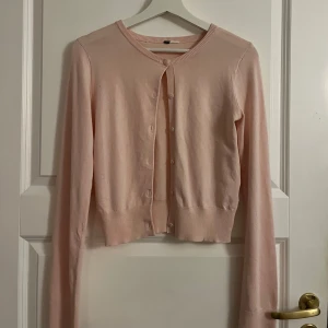 Ljusrosa kofta - Säljer en söt ljusrosa kofta med knappar framtill. Den är croppad och har långa ärmar, perfekt för lager-på-lager-stil. Passar bra till både jeans och kjol. Ett måste i garderoben för en stilren look!