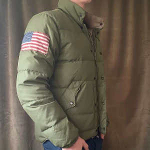 Grön dunjacka -  Ralph Lauren - Säljer en grön dunjacka från Denim & Supply Ralph Lauren med en amerikansk flagga på ärmen. Jackan har dragkedja och knappar framtill samt långa ärmar. Den har ett hål i ena fickan (se bild 5) men det går lätt att laga och är inget som märks vid användning. Vid minsta fråga eller fundering hör av er🤝