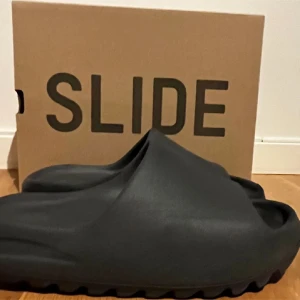 Svarta Yeezy Slide från Adidas - Säljer ett par svarta Yeezy Slide från Adidas. Dessa slides har en modern och minimalistisk design med en räfflad sula för extra grepp. Perfekta för en avslappnad stil.