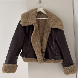 Brun skinnjacka med foder - Säljer en snygg brun skinnjacka med mysigt beige foder. Jackan har en krage och muddar i fuskpäls. Perfekt för höst och vinter, håller dig varm och stilren. Oversized i storlek. Går ner i pris vi snabb affär 