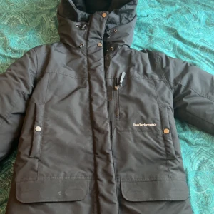 Svart dunjacka från Peak Performance - Säljer en svart dunjacka från Peak Performance med huva och flera praktiska fickor. Jackan har dragkedja och knappar framtill för extra skydd mot kyla. Perfekt för kalla vinterdagar.