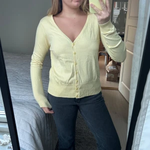 Gul kofta med knappar - Säljer en ljusgul kofta med knappar framtill. Den har en v-ringad design och långa ärmar, perfekt för vår och sommar. Koftan är i en mjuk och bekväm bomullsmix.