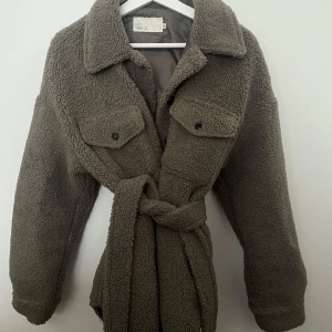 Mörkgrön teddyjacka från NLY Trend - Mysig  teddyjacka från NLY Trend med knappar och bälte i midjan. Jackan har två bröstfickor med knappar och en klassisk krage. Perfekt för kyliga dagar. Väldigt bra skick och bara använt några gånger en vinter.