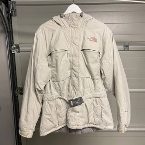 Beige jacka från The North Face - Säljer en beige jacka från The North Face i bra skick. Den har en praktisk huva och ett bälte i midjan för en snygg passform. Perfekt för kyliga höst- och vinterdagar. Jackan har långa ärmar och är gjord i ett slitstarkt material. Säljes i befintligt skick. Storlek: XL