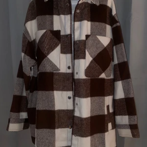 Brun och vit rutig overshirt - Snygg brun och vit rutig overshirt med knappar framtill. Perfekt för lager-på-lager-stil. Två stora bröstfickor ger en cool och praktisk touch. Passar perfekt för höst och vår.
