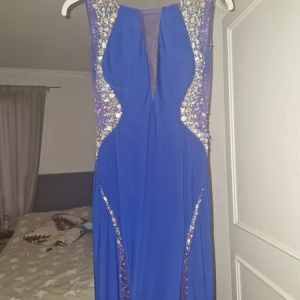 Blå långklänning med paljettdetaljer - Elegant blå långklänning med glittrande paljettdetaljer längs axlar och byst. Klänningen har en djup v-ringning och är ärmlös, vilket ger en stilren och festlig look. Perfekt för speciella tillfällen.