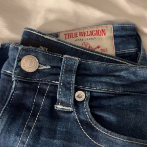 Blå jeans från True Religion - Säljer ett par snygga blå jeans från True Religion med klassisk femficksdesign och silverfärgade knappar. Bootcutmodell i storlek W26, sitter som en S.