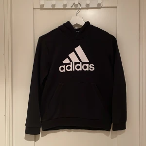 Svart hoodie från Adidas - Säljer en svart hoodie från Adidas i mycket bra skick. Den har en stor vit logga på framsidan och är perfekt för både vardag och träning. Hoodien har långa ärmar och en klassisk passform. Superbekväm och stilren!