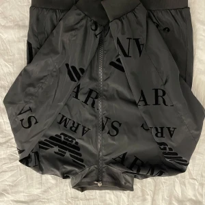 Svart bomberjacka från Emporio Armani - Snygg svart bomberjacka från Emporio Armani med stora logotyper och dragkedja framtill. Perfekt för en stilren look.