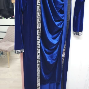 Blå långklänning med mönster - Elegant blå långklänning i sammet med grekiskt mönster längs kanterna och ett stort dekorativt motiv på ryggen. Klänningen har långa ärmar och en draperad framsida som ger en lyxig känsla.