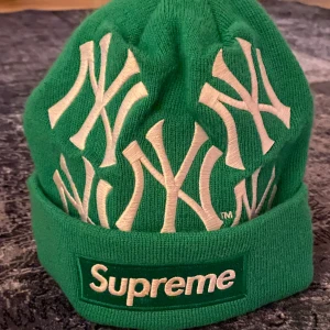 Grön mössa från Supreme - Snygg grön mössa från Supreme med broderade NY-loggor i vitt. Mössan har en uppvikt kant med Supreme-logga och texten 'World Famous' på baksidan. Perfekt för vintern 