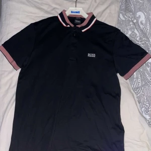 Svart pikétröja från Hugo Boss - Snygg svart pikétröja från Hugo Boss med röd och vit randig krage och ärmslut. Tröjan har en broderad logga på bröstet och är tillverkad i lyxig bomull för en bekväm passform.