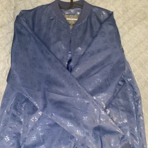 Blå bomberjacka från Louis Vuitton - Snygg blå bomberjacka från Louis Vuitton med ett diskret monogrammönster. Jackan har dragkedja och ribbade muddar vid hals och ärmslut. Perfekt för en stilren look. Knappt använd! Passar S/M. Pris kan diskuteras!