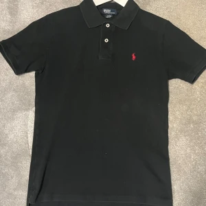 Svart pikétröja från Ralph Lauren för 14 åring - Snygg svart pikétröja från Ralph Lauren med klassisk krage och knappar. Den har en liten röd logga på bröstet som ger en stilren touch. Perfekt för en avslappnad men ändå stilfull look. 