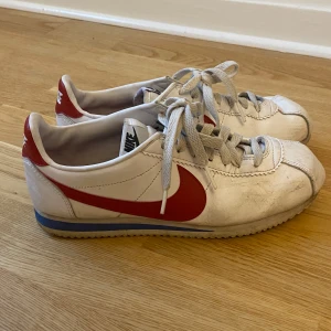 Nike Cortez i vitt och rött - Säljer ett par klassiska Nike Cortez sneakers i vitt med röd swoosh. De har en retro look med snörning och är i bra skick, men lite smutsiga. Perfekta för en casual stil eller träning. 