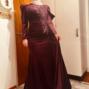 Vinröd långklänning med broderad detalj - Elegant vinröd långklänning i sammet med vackra broderade detaljer på framsidan. Klänningen har långa ärmar och en sofistikerad design som passar perfekt för festliga tillfällen. Den har en figurnära passform och en lyxig känsla.