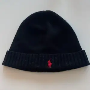 Snygg svart mössa från Polo Ralph Lauren i tvättbar merinoull. Den har en röd broderad logga framtill och ribbad kant. Perfekt för att hålla sig varm och stilren under kyliga dagar.   Tar emot bud.