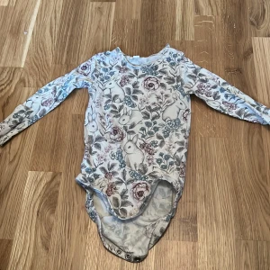 Blommig body från Newbie - Söt långärmad body från Newbie med ett charmigt blommönster och kaniner. Perfekt för de små under vår och sommar. Tryckknappar i grenen för enkel på- och avklädning.