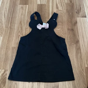 Svart klänning med rosett från H&M - Söt svart klänning från H&M med en ljusrosa rosett framtill. Klänningen har breda axelband och är ärmlös, perfekt för små barn. Disney-märke på etiketten.