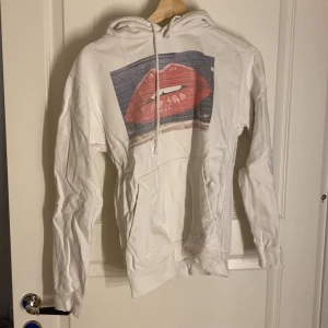 Vit hoodie med tryck av röda läppar - Säljer en vit hoodie med ett coolt tryck av röda läppar på framsidan. Hoodien har en klassisk design med dragsko i huvan och en stor magficka. 