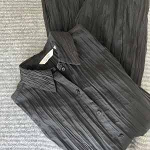 Svart plisserad set från & other stories  - Snygg svart plisserad set från & other stories. Skjortan har en klassisk krage och knappar framtill. Perfekt för en stilren look med en modern twist. Passar bra till både vardag och speciella tillfällen. Byxorna är utan fickor och raka i passform, det har mindre skada på sidorna. Lätt att sy ihop ( Se bild )