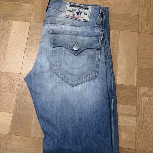 Blå jeans från True Religion - Säljer ett par snygga blå jeans från True Religion i straight fit. De har en cool slitning på framsidan och klassiska femfickor. Perfekta för en avslappnad stil. Passar bra till både vardag och fest! 