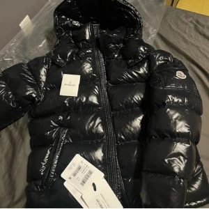 Svart pufferjacka från Moncler - Säljer en snygg svart pufferjacka från Moncler i mycket bra skick. Jackan har en glansig finish och är perfekt för kalla dagar. Den har en dragkedja framtill och en huva för extra värme. Passar perfekt för vintern och hösten!