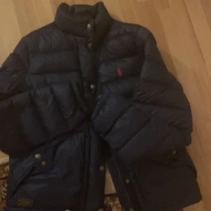 Svart dunjacka från Polo Ralph Lauren - Säljer en snygg svart dunjacka från Polo Ralph Lauren. Jackan har en klassisk design med röd broderad logga på bröstet och knappar framtill. Perfekt för kyliga dagar och håller dig varm och stilren. Passar både till vardags och lite finare tillfällen.