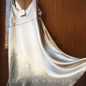 Silvrig klänning i Eco-SILK och Viskos - Elegant och glansig klänning i en vacker silvrig nyans. Den har smala axelband och knappar på sidan för en stilren look. Perfekt för festliga tillfällen!