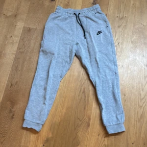 Grå mjukisbyxor från Nike - Säljer ett par grå mjukisbyxor från Nike med elastisk midja och snörning. Byxorna har en dragkedja på sidan och ribbade muddar vid bensluten. Perfekta för en avslappnad stil.