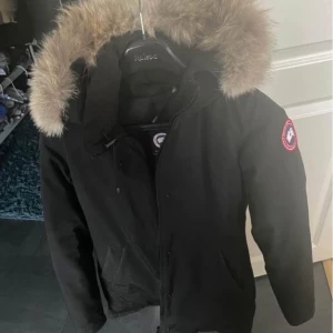 Svart parkajacka med päls från Canada Goose - Säljer en stilren svart parkajacka från Canada Goose med en mysig pälsfodrad huva. Jackan har en klassisk design med dragkedja och knappar framtill. Perfekt för kalla vinterdagar.
