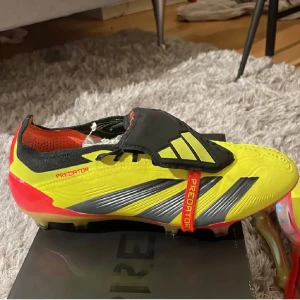 Adidas Predator fotbollsskor - Snygga Adidas Predator fotbollsskor i gult och rött med svarta detaljer. Skorna har en modern design med snörning och är perfekta för fotbollsplanen. De har en slitstark sula och är designade för optimal prestanda.