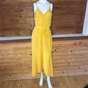 Gul långklänning med knytband - Snygg gul långklänning med smala axelband och v-ringning. Klänningen har ett knytband i midjan för en fin passform och ett diskret prickigt mönster som ger en extra touch. Perfekt för soliga dagar!
