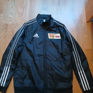 Svart träningsjacka FC Union Berlin. (Adidas) - Säljer en svart träningsjacka från Adidas med Union Berlin-logga och vita ränder på ärmarna. Jackan har dragkedja och är långärmad, perfekt för sportiga aktiviteter. Den har även ett tryck på baksidan som firar 100 år av Stadion An der Alten Försterei. Den passar folk som är mellan 160-175 cm