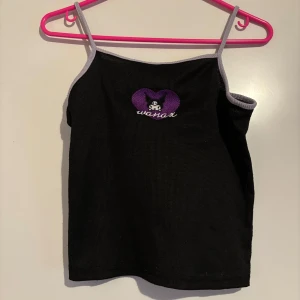 Svart linne med lila hjärta Kuromi - Säljer ett svart linne med smala axelband och en lila Kuromi hjärtapplikation med text framtill. Linnet har kontrasterande vita kanter som ger en snygg detalj. Perfekt för en avslappnad stil.