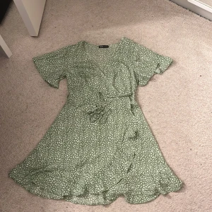 Grön kortklänning från Shein - Säljer en söt grön kortklänning från Shein med ett vitt mönster. Klänningen har korta ärmar och en omlott-design med knytning i midjan. Perfekt för en somrig look! 🌿