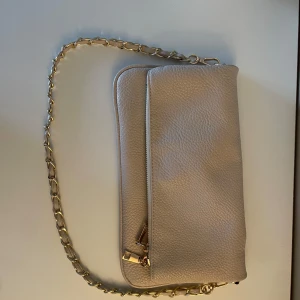 Beige axelväska med kedja - Snygg beige axelväska med guldfärgad kedja som axelrem. Väskan har en dragkedja och magnetknappar för säker förslutning. Perfekt för att bära med sig det viktigaste med stil.
