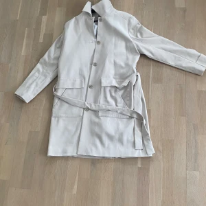 Beige trenchcoat från Stockh LM - Snygg beige trenchcoat från Stockh LM i storlek 44. Jackan har långa ärmar och ett bälte i midjan för en stilren look. Perfekt för vår och höst med sina klassiska knappar och fickor framtill.