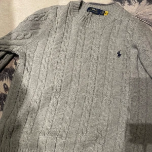 Grå stickad tröja från Polo Ralph Lauren storlek S - Säljer en stilren grå stickad tröja från Polo Ralph Lauren. Tröjan har ett klassiskt flätmönster och en broderad logotyp på bröstet. Perfekt för kyligare dagar med sin mjuka och bekväma passform.