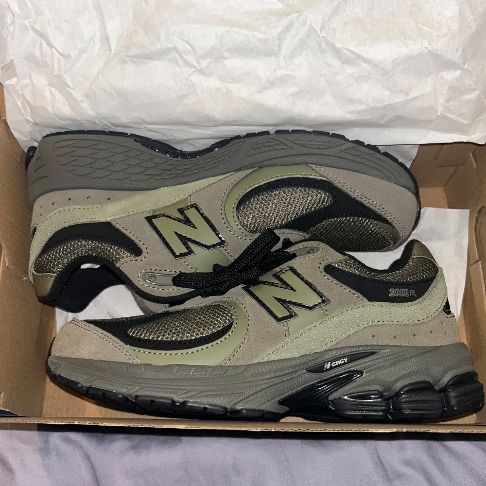 Snygga New Balance 2002R sneakers i en kombination av grått och olivgrönt. Skorna har en robust sula och är tillverkade i en mix av syntet och mocka. Perfekta för en casual look med sin klassiska snörning och bekväma passform.. Kengät.
