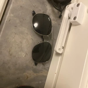 Ray-Ban solglasögon  - Snygga svarta solglasögon från Ray-Ban med klassisk design. De har en stilren metallram och mörka linser. Perfekta för att ge en cool touch till din stil. Kommer med ett svart fodral för säker förvaring.