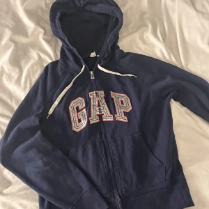 Mörkblå hoodie från GAP - Säljer en mörkblå hoodie från GAP med dragkedja och stor logga på bröstet. Tröjan har en justerbar huva med vita snören och praktiska fickor framtill. 
