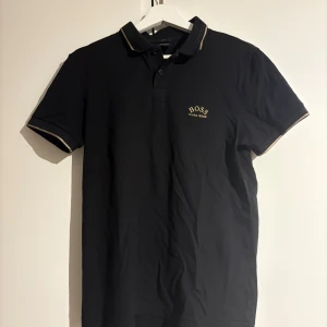 Svart pikétröja från BOSS - Snygg svart pikétröja från BOSS med gulddetaljer på kragen och ärmsluten. Perfekt för en stilren look. Tröjan har korta ärmar och en klassisk krage med knappar.