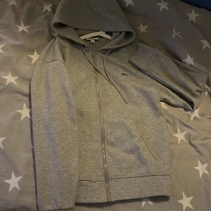 Grå hoodie från Lacoste  - Säljer en klassisk grå hoodie från Lacoste med dragkedja och huva. Den har en liten krokodil-logga på bröstet och är perfekt för en avslappnad stil. Passar bra till både jeans och joggers.