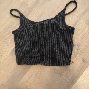 Svart glittrig linne - Snygg svart linne med glittriga ränder. Den är ärmlös med smala axelband och har en croppad passform. Perfekt för en kväll ute eller en festlig tillställning.