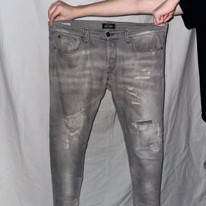 Grå jeansbyxor från jack m Jones  - Snygga grå jeansbyxor från  jack b Jones med en sliten look och skinny passform. Byxorna har en klassisk femficksdesign och är perfekta för en trendig stil. De har en knapp och dragkedja framtill. 33/32