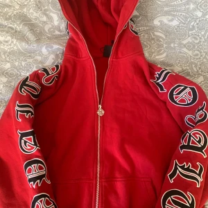 Röd hoodie med tryck - Snygg röd hoodie med dragkedja och tryck på ärmarna. Den har en stor huva och en cool design med text i svart och vitt. Perfekt för en avslappnad stil.