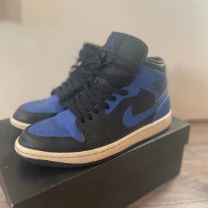 Nike Air Jordan 1 Mid i svart och blå - Snygga Nike Air Jordan 1 Mid sneakers i en cool kombination av svart och blå. Skorna har klassisk snörning och en ikonisk design som passar perfekt för streetwear-stilen.
